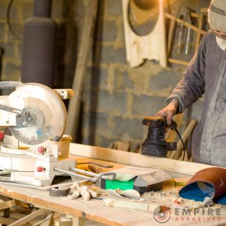 Woodworker using an electric sander on wood in a workshop, with Empire Abrasives logo, illustrating proper sanding technique for paint preparation